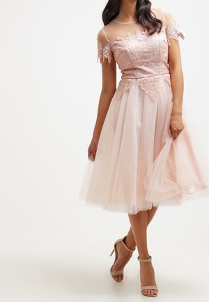 Femme portant une robe top en dentelle rose clair avec des manches transparentes et une jupe en tulle fluide, associée à des talons beige à bride à la cheville.