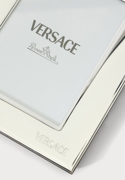 Cadre photo en métal argenté avec le logo Versace gravé sur le bord inférieur et texte imprimé à l'intérieur du cadre.