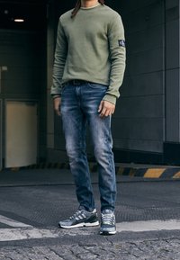 Olivgrön långärmad tröja, blå jeans och grå sportskor med gröna detaljer. Har logopatch på ärmen och en åtsittande design.