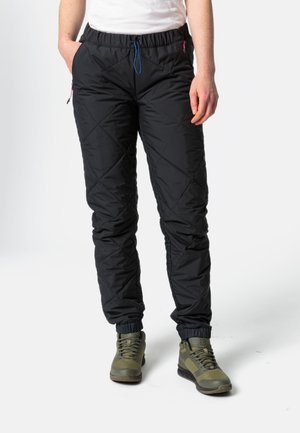 Gequiltte zwarte broek met een elastische tailleband, blauwe trekkoord en zijzakken. De boorden gatheren bij de enkels. Gecombineerd met groene wandelschoenen.