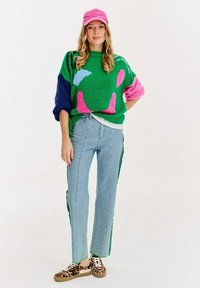 Mehrfarbiger gestrickter Pullover in grün mit pinken und blauen Mustern, kombiniert mit hellblauen Jeans mit grünen Seitenstreifen und Schuhen im Leopardenmuster.