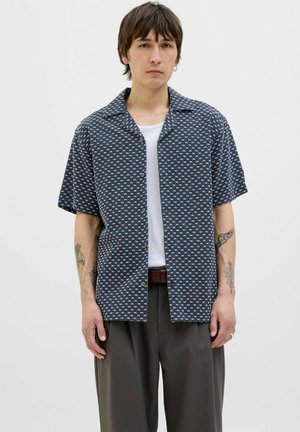 Chemise à manches courtes en bleu marine avec un motif géométrique blanc, design à boutons, texture douce et coupe décontractée, portée sur un t-shirt blanc.