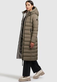 Long, olive-green puffer coat with a hood, featuring boxy quilted sections, zippered front, and drawstring accents. Paired with light-colored platform shoes.
