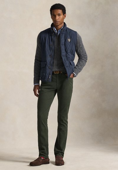 Μπλε σκούφο γιλέκο με καπιτονέ, γκρι πουλόβερ με πλέξη καλώδιο, πράσινο παντελόνι, καφέ δερμάτινα παπούτσια σουέτ με κόκκινες λεπτομέρειες, σχεδιασμός με στρώσεις και εφαρμοστή γραμμή.