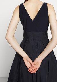 Henkilö, joka on pukeutunut hihattomaan mustaan mekkoon, jossa on V-muoto takana, kädet yhteen pantuna selän taakse, näkyy selkäpuolelta yksinkertaista taustaa vasten.