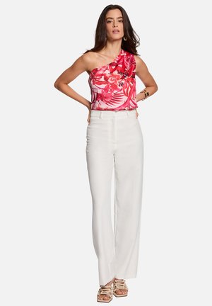Femme portant un top floral rose asymétrique, un pantalon blanc taille haute à jambes larges, et des sandales à talons beige à brides, posant les mains sur les hanches.