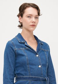 Τζάκετ τζιν με γιακά, κάθετους ραφές, μπροστινά κουμπιά και ζώνη. Μπλε χρώμα με λεία, υφασμάτινη υφή.