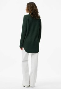 Maglione a maniche lunghe di colore verde scuro, con orlo asimmetrico, tessuto a trama, e spalle ribassate, abbinato a pantaloni bianchi a gamba larga.