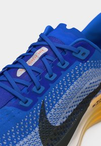 Κοντινό πλάνο μπλε παπουτσιού τρεξίματος Nike με μαύρο λογότυπο, πλεκτό επάνω μέρος, μπλε κορδόνια και πορτοκαλί επένδυση στη σόλα.