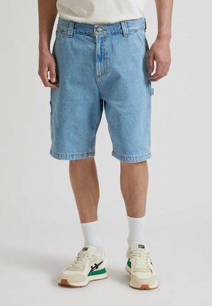 Pantalones cortos vaqueros azul claro con un corte relajado, cinco bolsillos y detalles de costura. Combinados con calcetines blancos y zapatillas beige.