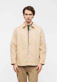 Μπεζ βαμβακερό μπουφάν με κούμπωμα με κουμπιά, δύο μπροστινές τσέπες και διακριτικό λογότυπο. Το μοντέλο φοράει πράσινο πουκάμισο από κάτω.