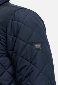 Giacca trapuntata di colore blu scuro con una texture liscia, caratterizzata da motivi a rombi e un'etichetta nera con "EST.1993" sulla spalla.