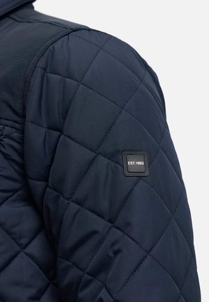 Gequilted donkerblauwe jas met een gladde textuur, voorzien van ruitpatronen en een zwart label met "EST.1993" op de schouder.