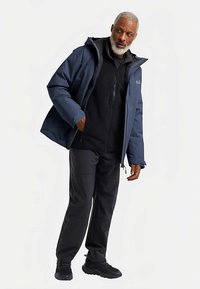 Waterafstotend donkerblauw jack met capuchon, gecombineerd met een zwarte fleece met rits en zwarte broek. Heeft een ontspannen pasvorm en duurzaam materiaal.