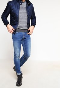 Mies, joka pukeutuu tummansiniseen tikattuun takkiin harmaapilkullisen neuleen, sinisten farkkujen ja mustien nauhakenkien kanssa, seisoo jalat ristissä.