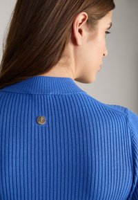 Blauer gerippter Strickpullover mit hohem Kragen und einem runden Logo-Metallakzent auf der Rückseite. Zeigt die Textur und die definierten Linien des Stoffes.