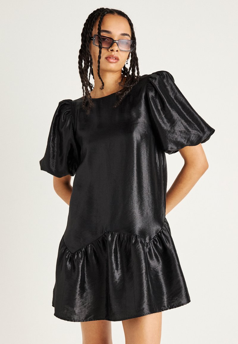 Μαύρο φόρεμα από σατέν με μανίκια puff και φαρδύ τελείωμα. Λείο υλικό, στρογγυλή λαιμόκοψη και δομημένοι ώμοι δημιουργούν όγκο στη σιλουέτα.