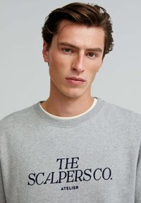 Gray sweatshirt with round neck and ribbed cuffs, featuring navy blue embroidered text "THE SCALPERS CO. ATELIER" on the front.