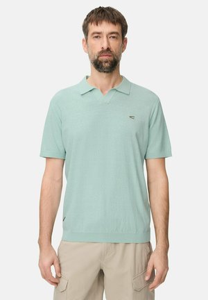 Mężczyzna z krótkimi brązowymi włosami i brodą, ubrany w jasnooliwkową koszulkę polo z krótkim rękawem oraz beżowe krótkie spodnie typu cargo, stoi na tle jednolitego tła.