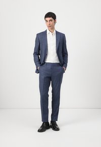 Jūrinės mėlynos spalvos kostiumas, kurio sudėtyje yra pritaikyta švarkas ir siauri kelnių, pagamintas iš tekstūruoto audinio. Derinamas su balta marškiniais ir juodais batais.