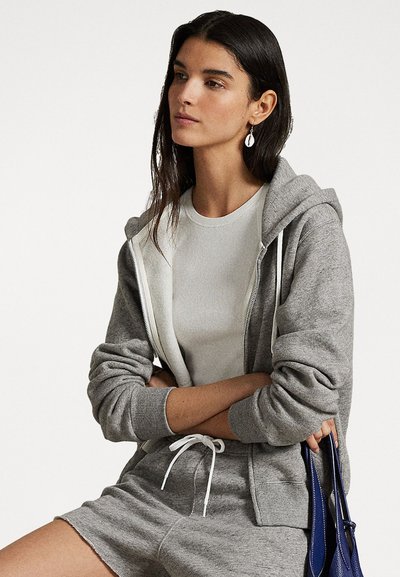 Grijze hoodie met een voorrits en een trekkoord, gedragen over een witte getailleerde T-shirt, gecombineerd met grijze shorts en een blauwe handtas.
