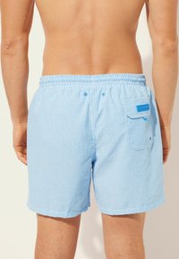 Světle modré plavky s pruhy, elastickým pasem, zadní kapsou a dvěma knoflíky. Měkká textura a délka do poloviny stehna.