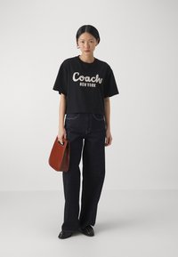 Zwarte cropped T-shirt met "Coach New York" in crème bedrukt, gecombineerd met wijde donkere jeans en een kleine bruine handtas.
