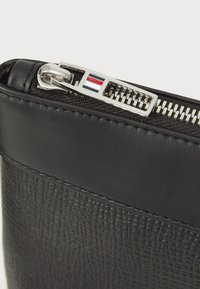 Sac en cuir noir avec un panneau texturé, une section supérieure lisse et un tirette en métal argenté avec des accents rouge et bleu.