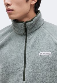 Άνδρας φοράει ανοιχτό γκρι fleece μπουφάν με ψηλό γιακά και φερμουάρ μπροστά, με λογότυπο Columbia στο αριστερό στήθος.