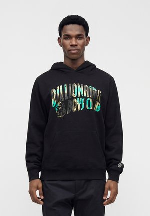 Jeune homme portant un sweat à capuche noir avec le texte « Billionaire Boys Club » en dégradé bleu et beige, debout devant un fond clair uni.