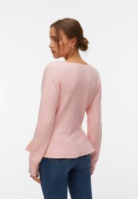 Femme aux cheveux bruns coiffés en chignon, portant un pull rose pâle à manches longues et un jean bleu, légèrement tournée sur le côté sur un fond blanc.