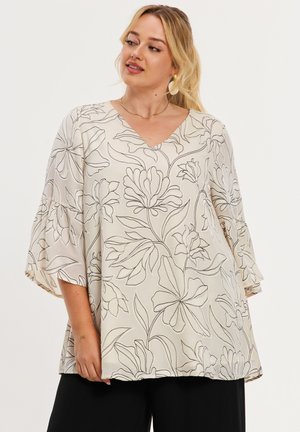 Kvinde iført en løs cremefarvet bluse med sorte blomsterkonturer og brede trekvartærmer, kombineret med sorte bukser.