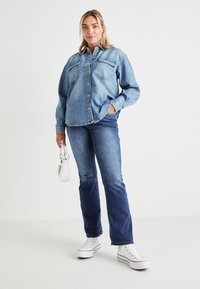 Camisa de mezclilla azul claro con dos bolsillos en el pecho, cierre de botón y mangas largas, combinada con jeans azul oscuro y zapatillas blancas.