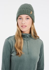 Beanie en tricot vert forêt avec un motif texturé et une petite étiquette de marque, associé à un haut zippé assorti avec un design en tricot torsadé.