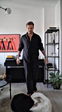 Uomo in camicia nera e pantaloni neri sta a piedi nudi nel soggiorno con pianoforte nero, opere d'arte astratta, scaffale in metallo e due cani che si rotolano sul pavimento.