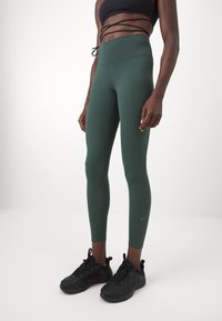 Leggings de cintura alta, de color verde oscuro, hechos de un material elástico. Presentan una textura suave y un sutil logo de Nike en la pierna.