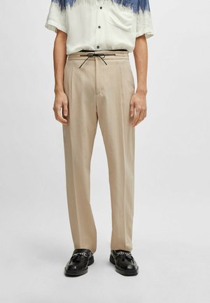 Beige Hosen in lockerer Passform mit einem Kordelzugbund und glatter Textur. Kombiniert mit schwarzen Schuhen und einem mehrfarbigen Hemd.