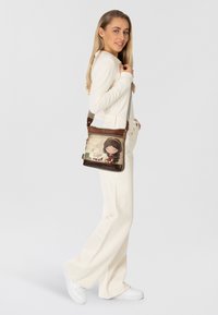 Braune Lederhandtasche mit einer verspielten Illustration und Text auf der Vorderseite, verstellbarem Riemen und Reißverschlussfächern.