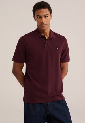 Homme portant un polo bordeaux foncé à manches courtes et un jean bleu foncé, debout les mains dans le dos devant un fond uni.