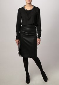 Zwarte blouse met lange mouwen en geplooide halslijn, versierd met embellishments, gecombineerd met een getailleerde zwarte leren kokerrok. Zwarte panty’s en hakken.