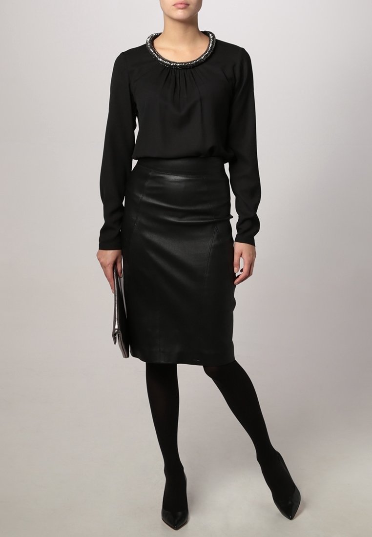 Zwarte blouse met lange mouwen en geplooide halslijn, versierd met embellishments, gecombineerd met een getailleerde zwarte leren kokerrok. Zwarte panty’s en hakken.