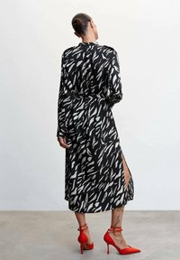 Robe à motifs noir et blanc avec des manches longues, taille cintrée et fente latérale ; assortie à des chaussures à talons hauts rouges vives.