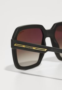 Ochelari de soare negri, supradimensionați, cu lentile gradient, având un finisaj mat, o formă unghiulară îndrăzneață și un accent auriu pe tâmplă, cu branding „CARRERA”.