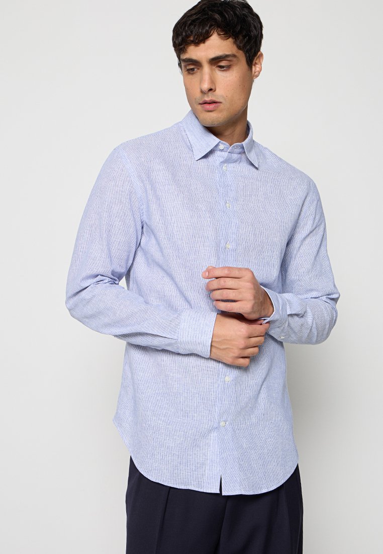 Hombre ajustándose el puño de la camisa azul clara texturizada de manga larga con botones, con pantalones oscuros, sobre un fondo claro y liso.