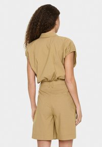 Lühikeste varrukatega khaki jumpsuit, millel on vormitud vöökoht, elastne riba ja suured küljetaskud. Sile tekstuur, lõdva disainiga ja kohandatud kuju.