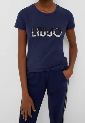 Kvinde iført marineblå t-shirt med skinnende "LIU JO" tekst og matchende marineblå bukser med sølvfarvede striber på siden, hånd i lommen.