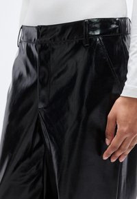 Μαύρο δερμάτινο παντελόνι με γυαλιστερό φινίρισμα, με ραμμένη σχεδίαση, πιέτες μπροστά και πλαϊνές τσέπες. Κοντινή λήψη που αναδεικνύει την υφή.