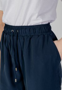 Pantaloni corti di colore blu navy con vita elasticizzata, cordoncino con estremità metalliche, tasche laterali e una texture di tessuto liscia e leggera.