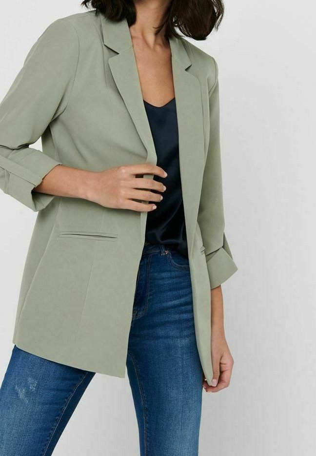 Femme portant un blazer vert clair par-dessus un top noir avec un jean bleu sur un fond clair uni.