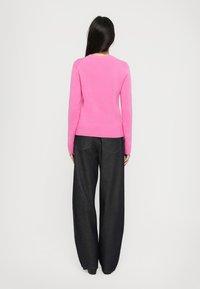 Frau mit langen, dunklen Haaren, die einen leuchtend pinken Pullover und locker sitzende, dunkelblaue Jeans trägt, steht vor einem weißen Hintergrund.